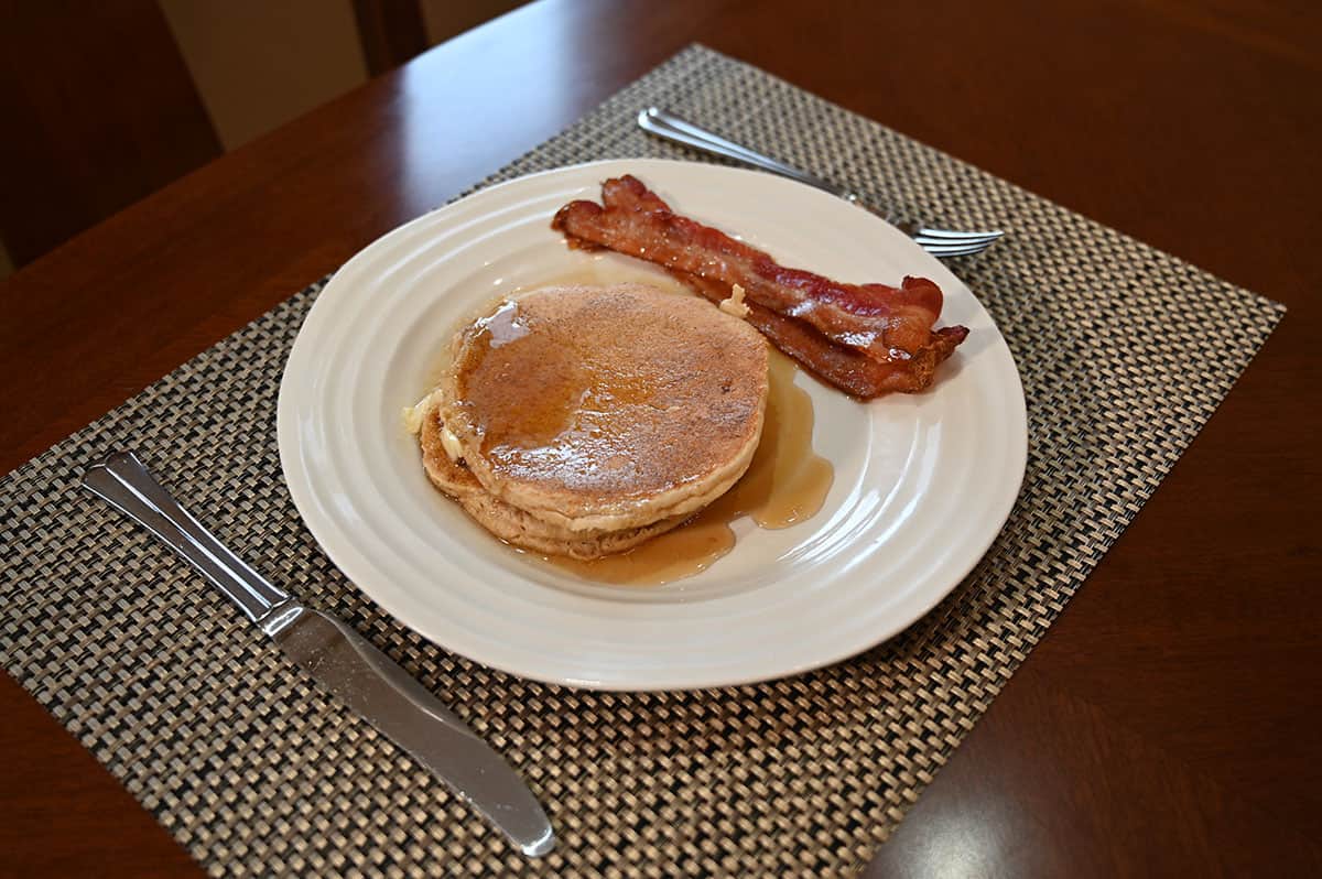 Side view image of a white plate with two pancakes on it and two strips of bacon with a fork and knife beside the plate.