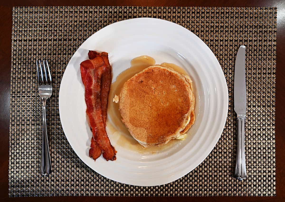 Top down image of a white plate with two pancakes on it and two strips of bacon with a fork and knife beside the plate.