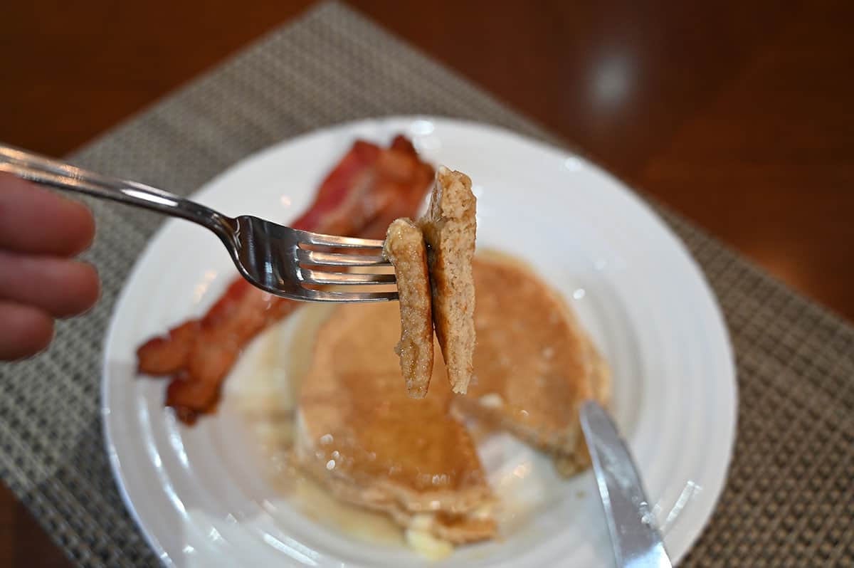 Closeup image of a forkful of Kodiak Power Cakes with a plate in the background with pancakes and bacon on the plate.