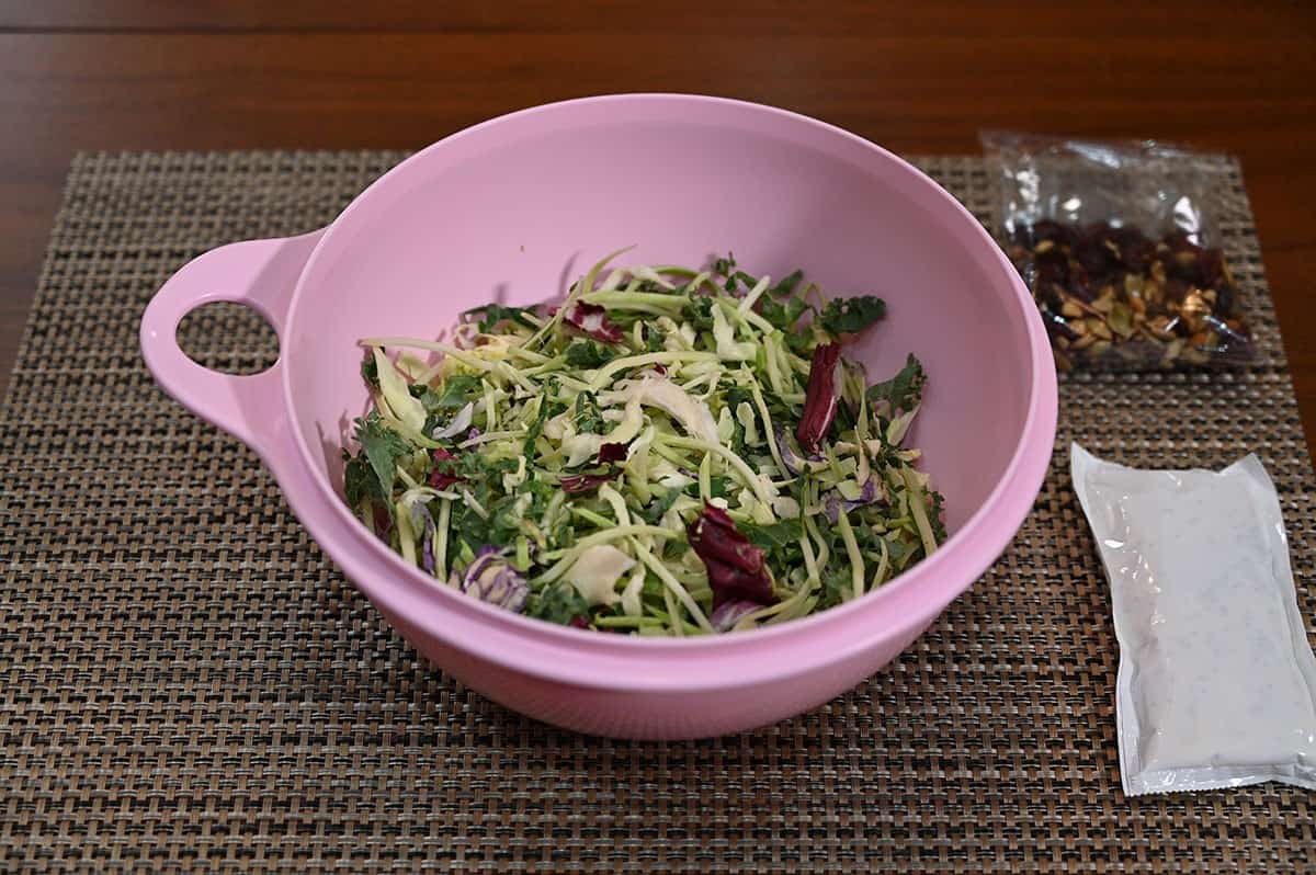 Costco Eat Smart Sweet Kale Salad Kit poured into a pink bowl with cranberries, pumpkin seeds and dressing beside the bowl. 