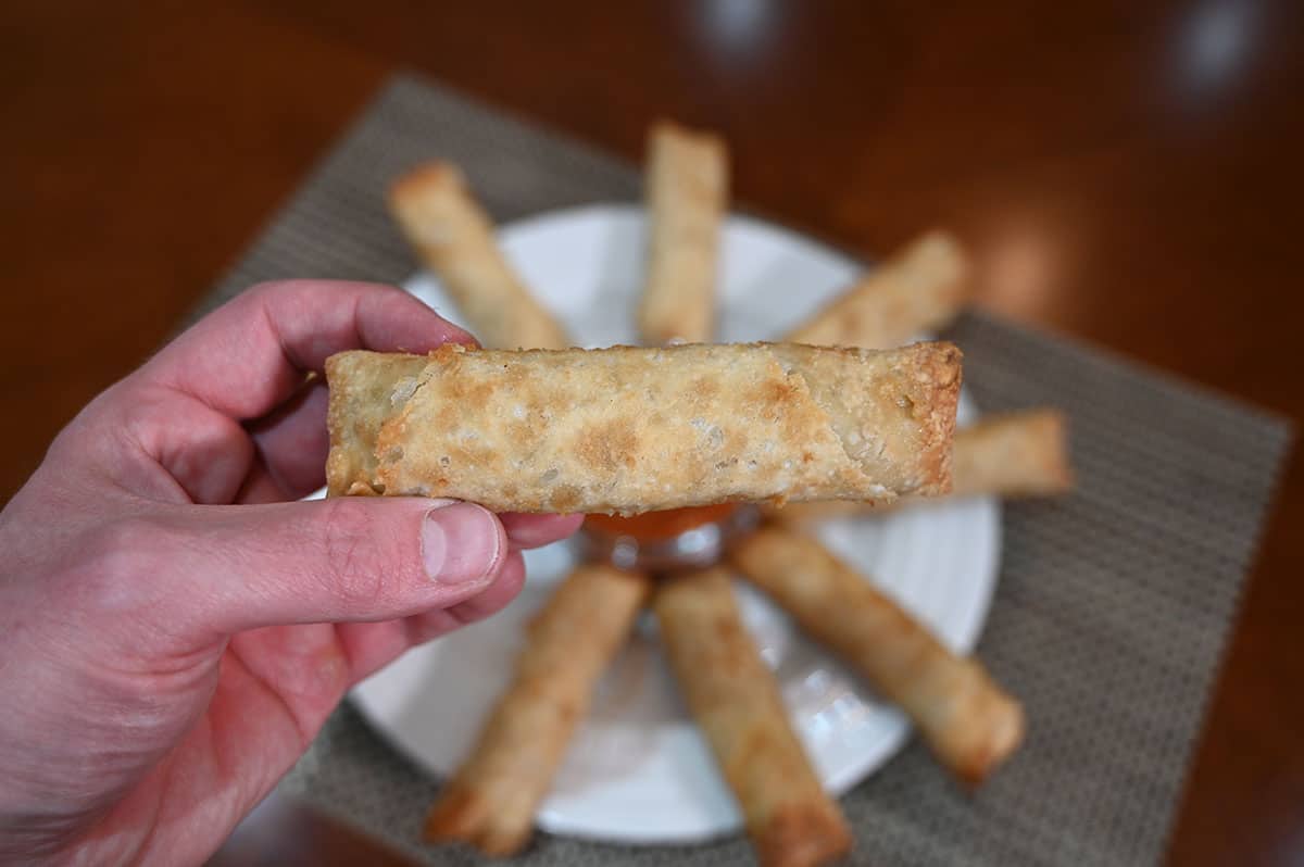 Closeup image of a hand holding one egg roll with a plate of egg rolls in the background.