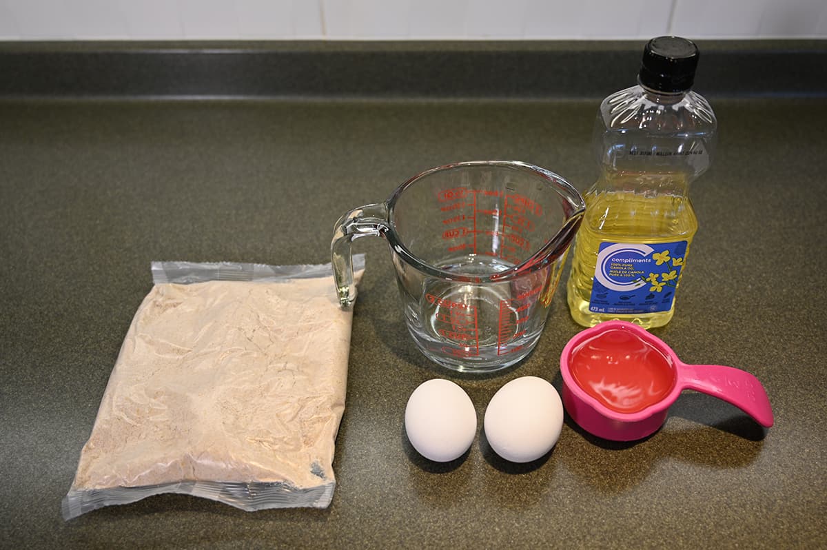 Top down image of a bag of mix beside two eggs, canola oil, and a measuring cup with water in it.