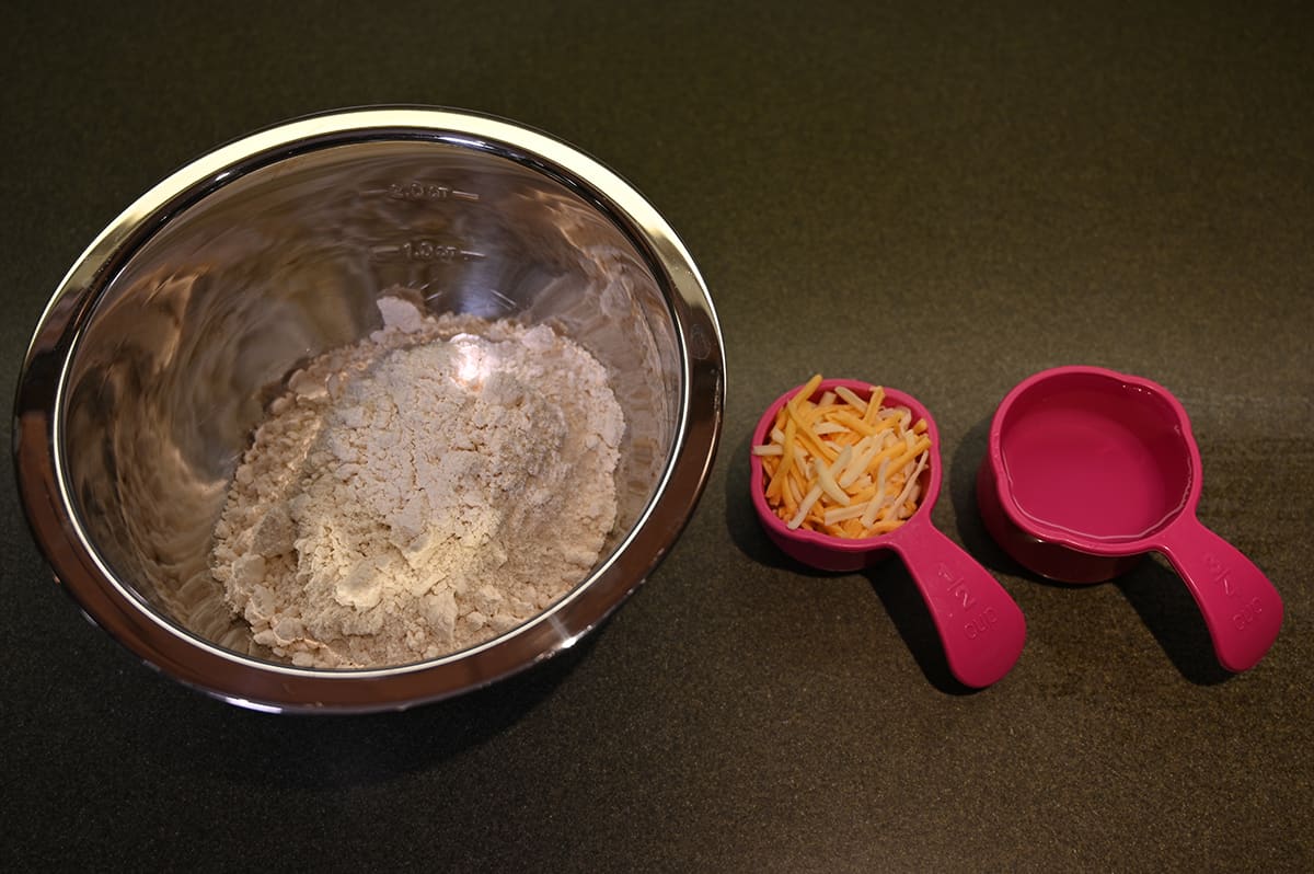 Image of biscuit mix poured into a metal bowl with a measuring cup of water and a measuring cup of cheese beside the metal bowl.