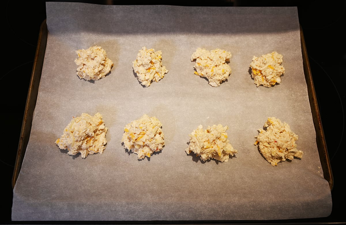 Image of a baking tray lined with parchment paper sitting on top of an oven with eight unbaked biscuits on the tray.