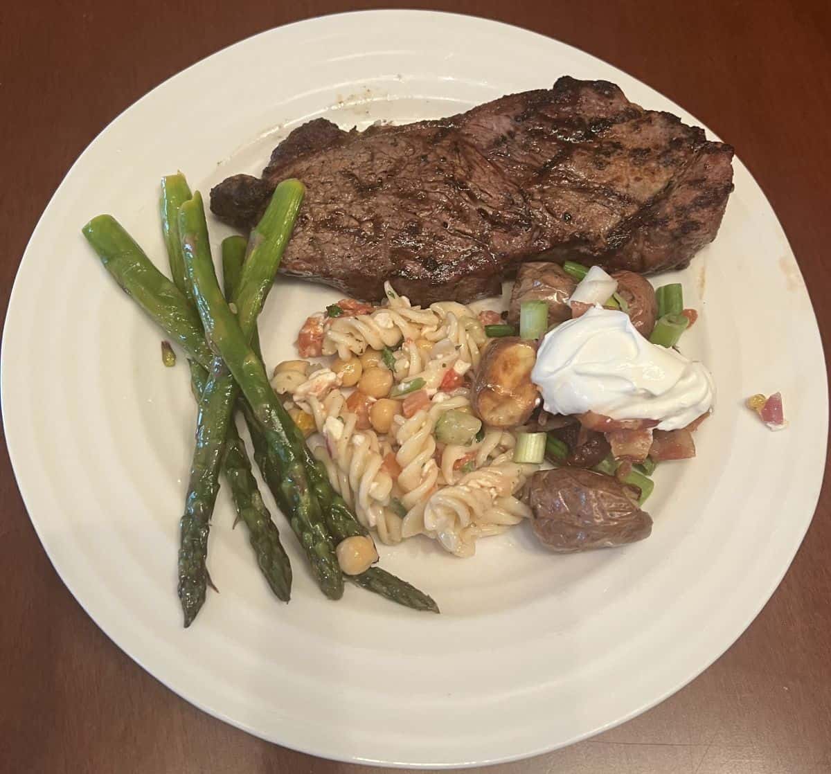 Top down image of a plate with steak on it, asparagus, potatoes and pasta salad.
