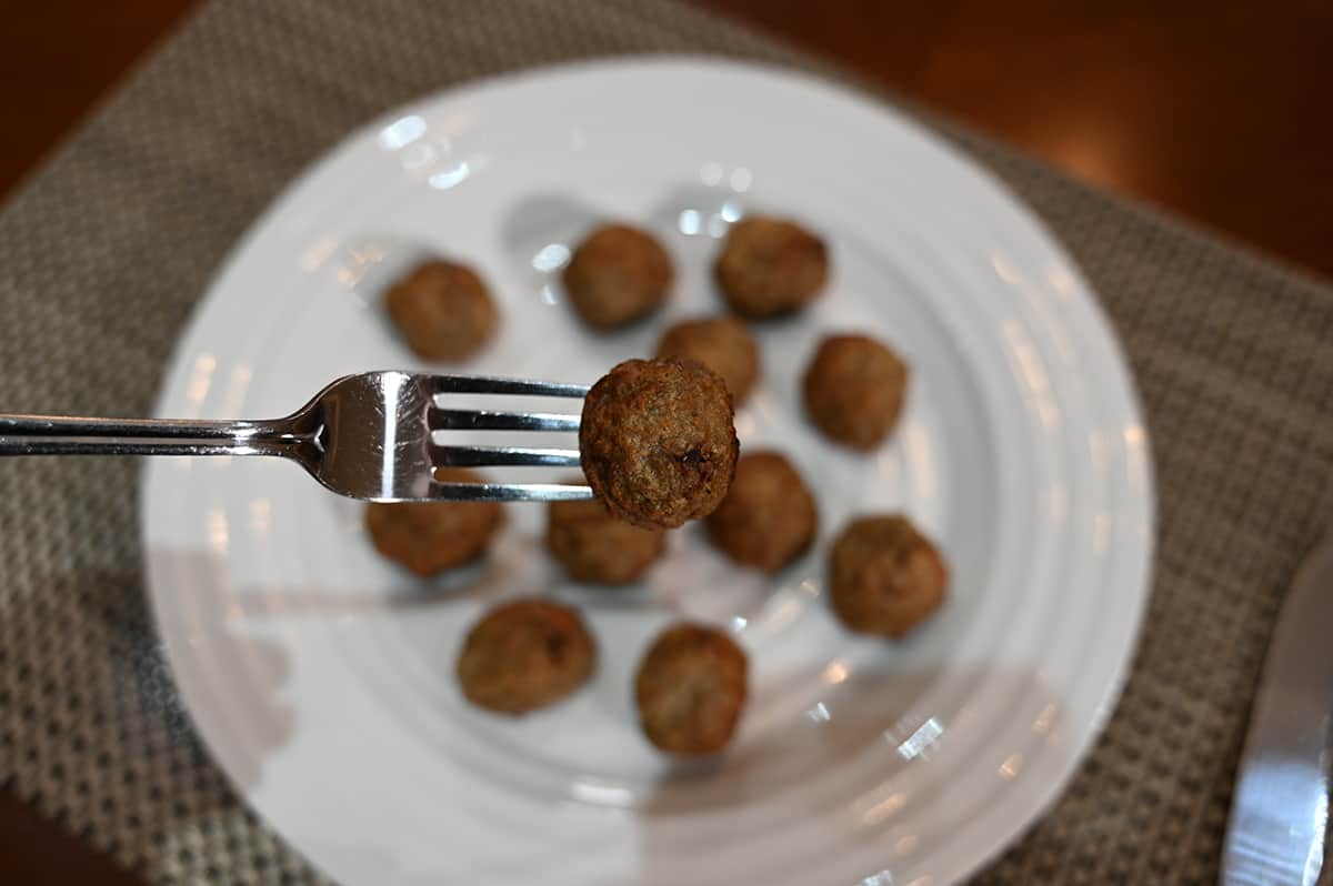 Closeup image of a fork with one meatball on it and a plate of cooked meatballs in the background.