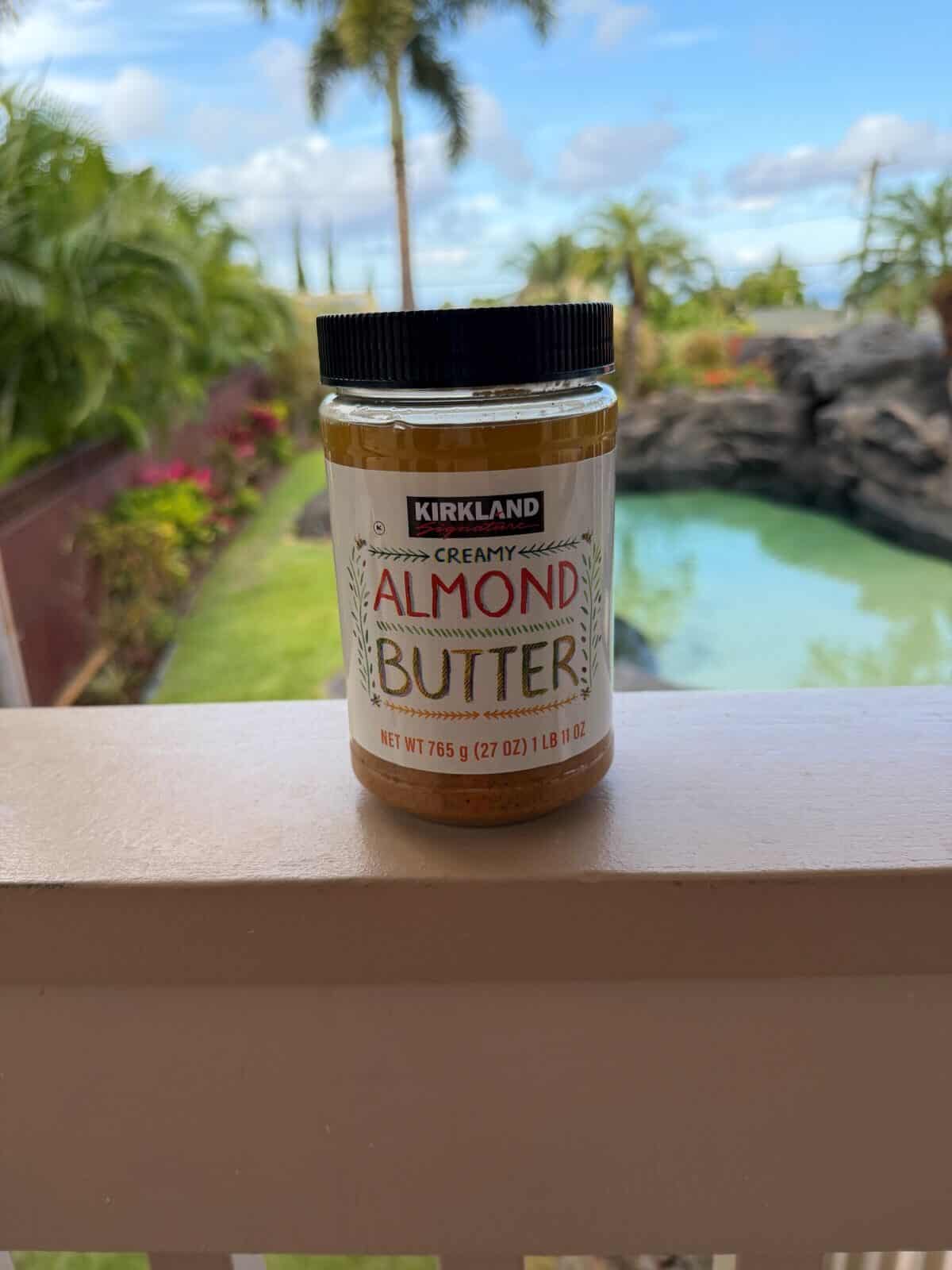 Image of an unopened jar of the Costco Kirkland Signature Creamy Almond Butter sitting on a deck outside with a pool in the background.