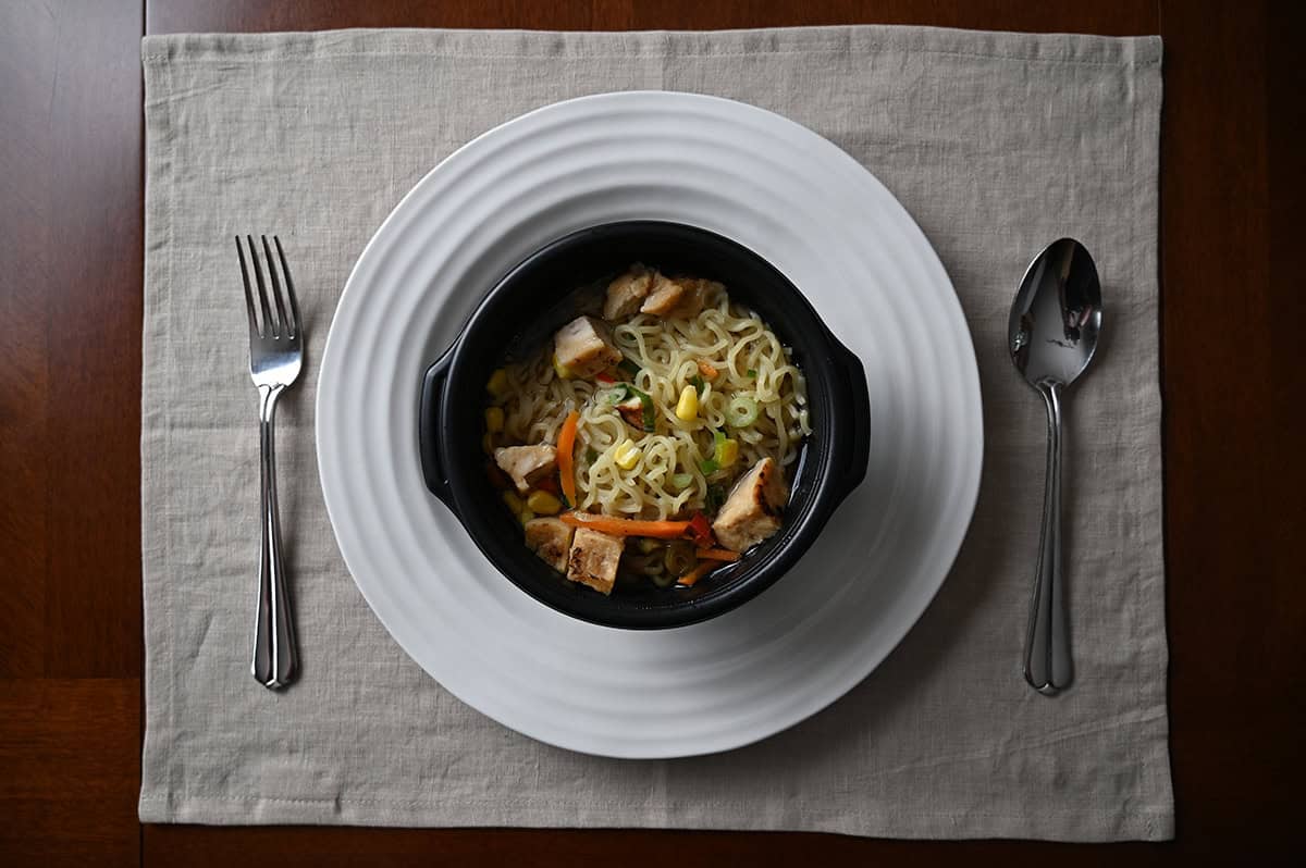 Top down image of a prepared bowl of ramen, served on a white plate beside a fork and spoon.