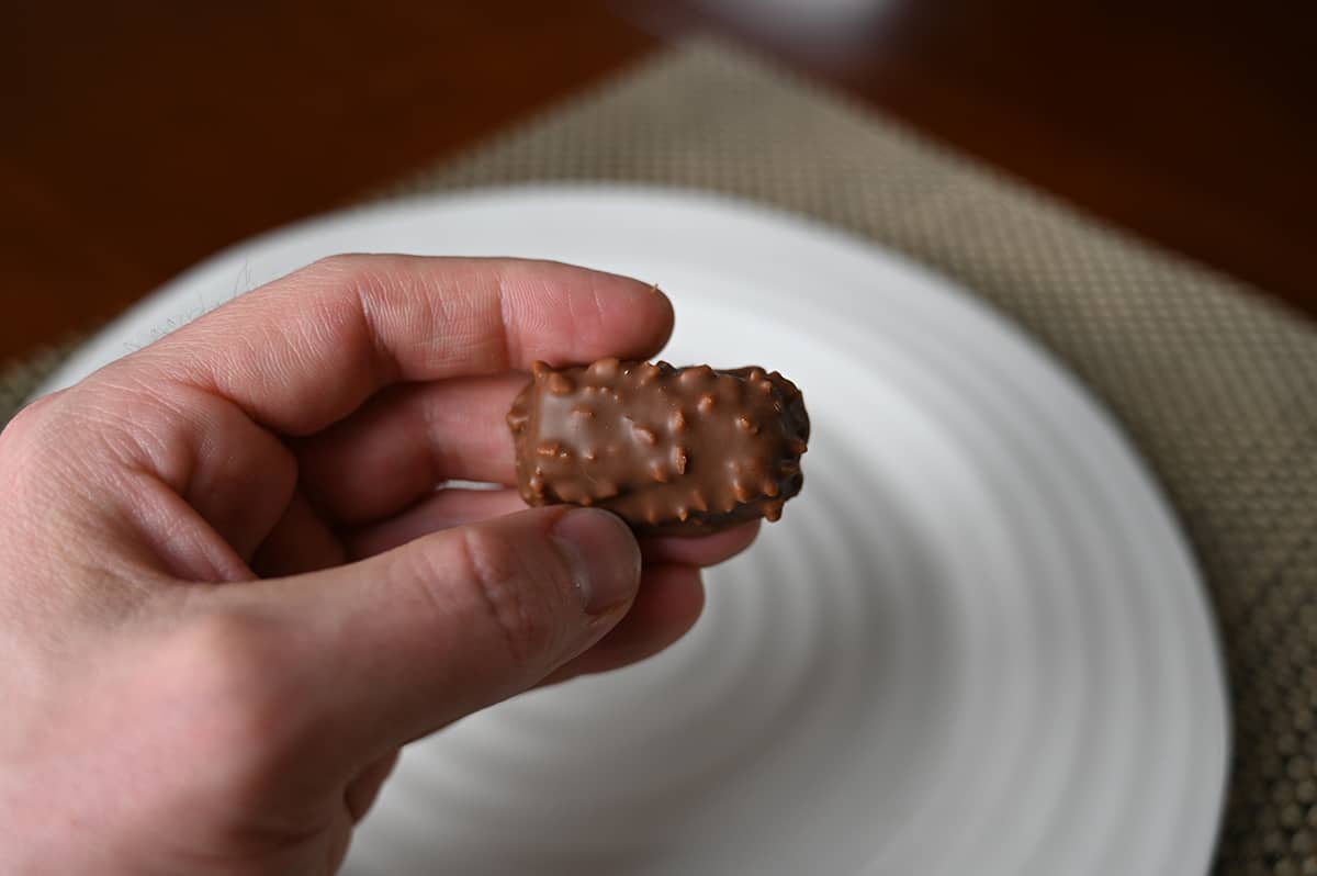 Image of a hand holding one crunchy praline chocolate close to the camera with a plate of chocolates in the background.