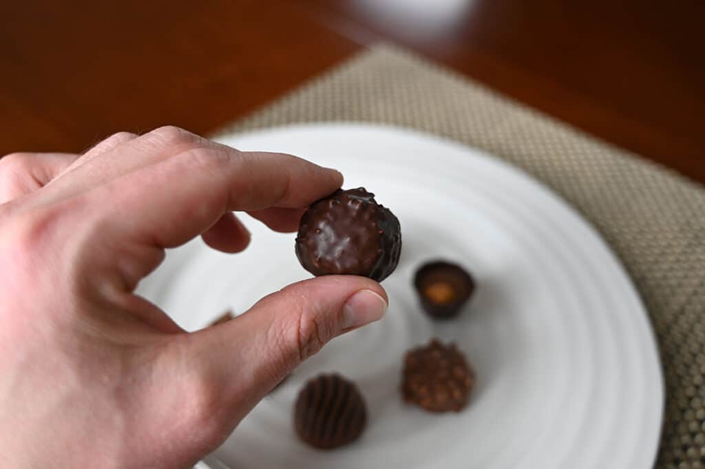 Image of a hand holding one dark cluster chocolate close to the camera with a plate of chocolates in the background.
