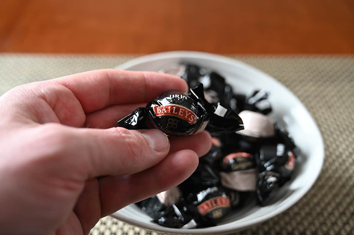 Image of a hand holding one wrapped Baileys chocolate close to the camera with a bowl of chocolates in the background.