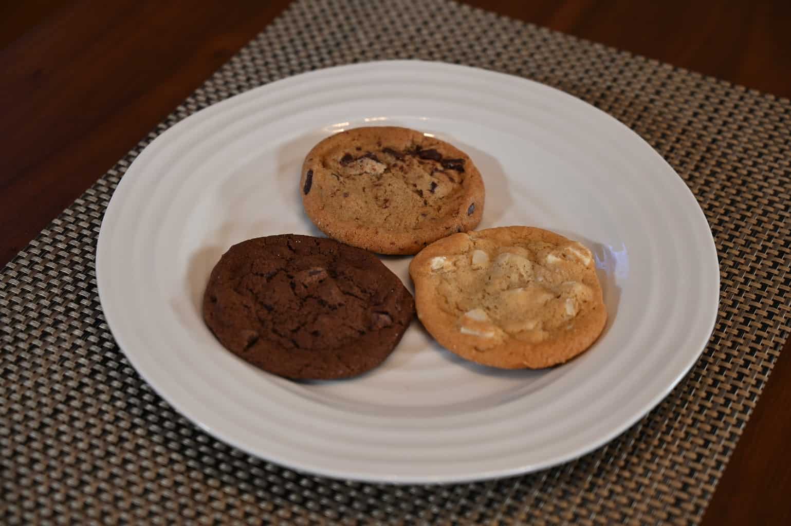 A white chocolate macadamia nut cookie, a triple chocolate cookie and a chocolate chunk cookie on a plate photographed from the side.