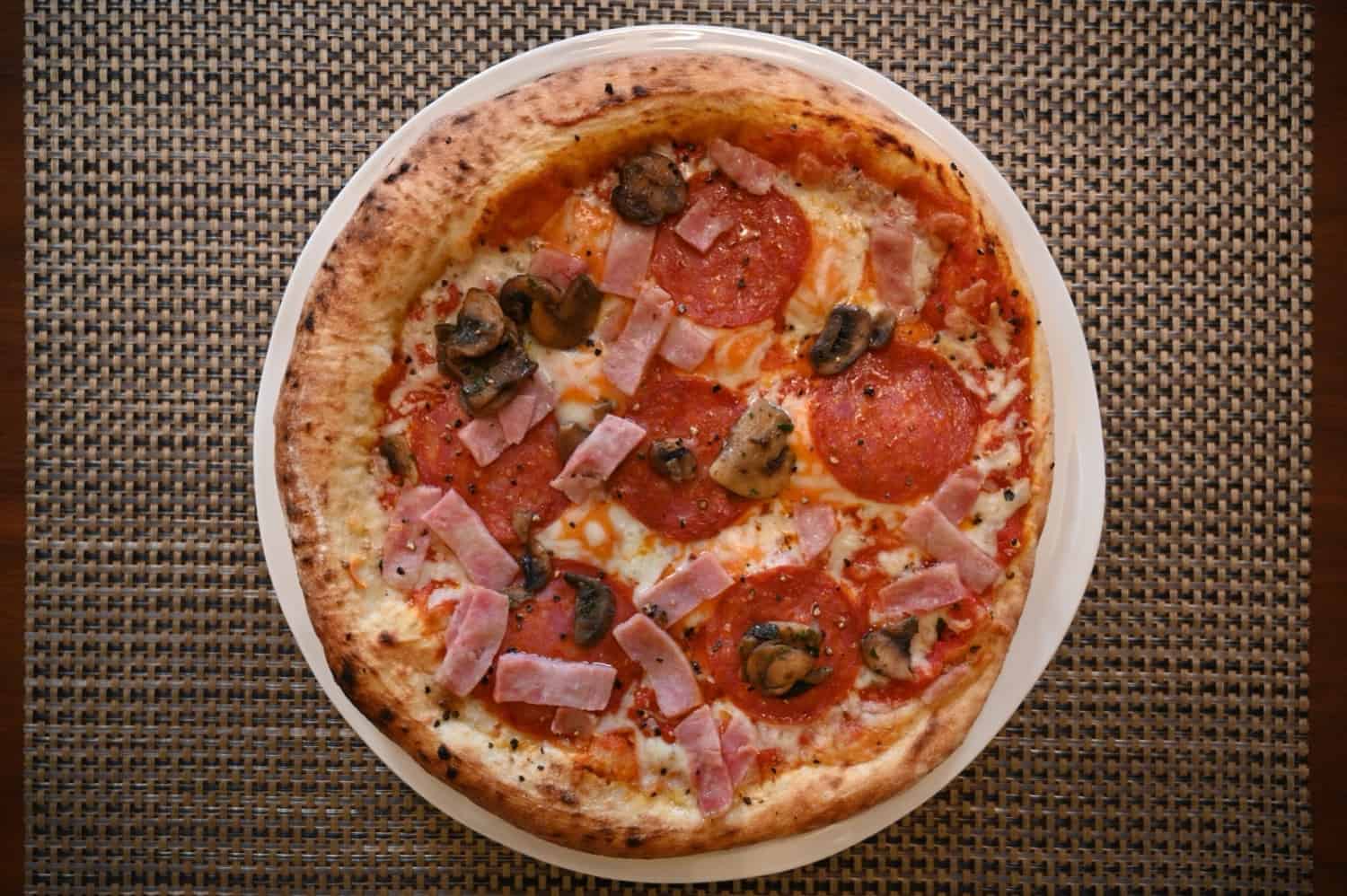 Top down image of a salami, mushroom and bacon pizza cooked and served on a white plate.