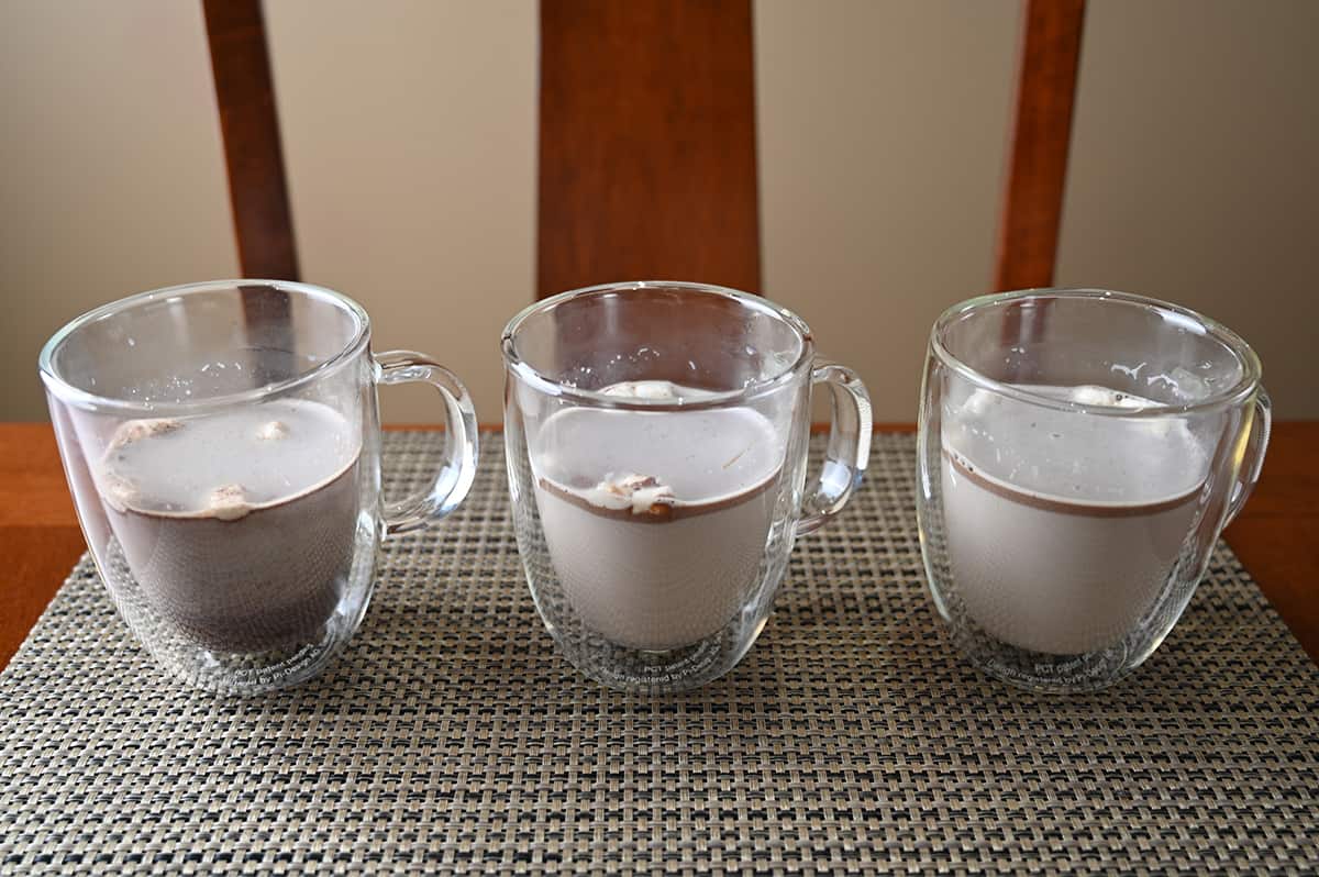 Image of three clear mugs with milk and hot chocolate bombs mixed in after mixing them in with a spoon. There are marshmallows floating on the top.