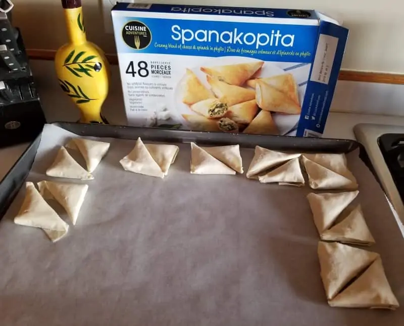 Photo of a tray of uncooked spanakopita in front of the Cuisine Adventures Spanakopita box.