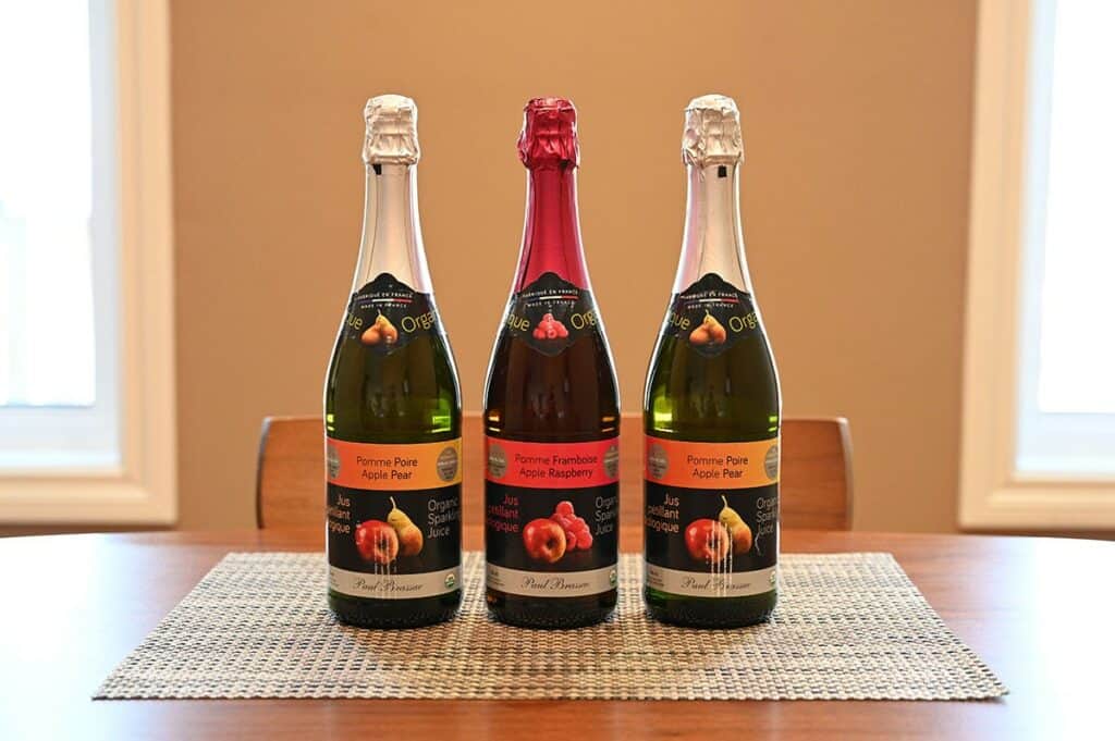 Image of the Costco Paul Brassac Organic Sparkling Juice three bottles, apple pear, apple raspberry and apple pear sitting on a table unopened. Side view image. 