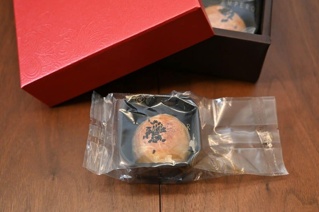 One individually wrapped Costco Emperor Lava Pastry with Lotus Seed Paste in wrapper sitting on brown table. 