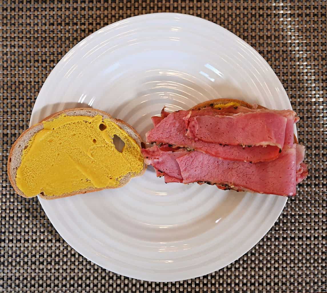 Open faced Schwartz's Smoked Meat sandwich with mustard on one slice of bread and meat stacked on the other piece of bread. On a white plate. 
