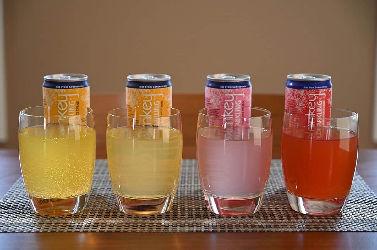 Image of the four different kinds of sparkling juice drink poured into glasses sitting on a table with the can of the flavor poured behind each glass. 