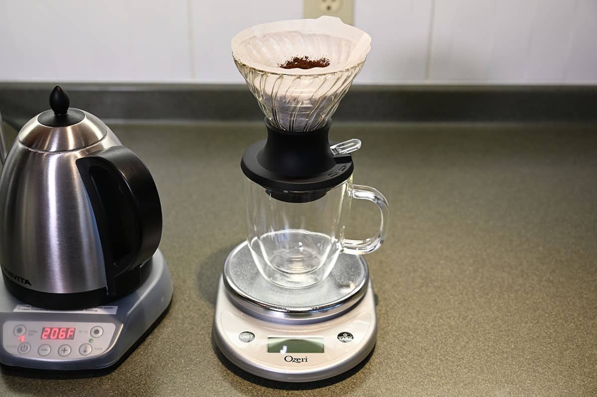 Image of coffee equipment on a counter, a kitchen scale, kettle, coffee cup and filter. 