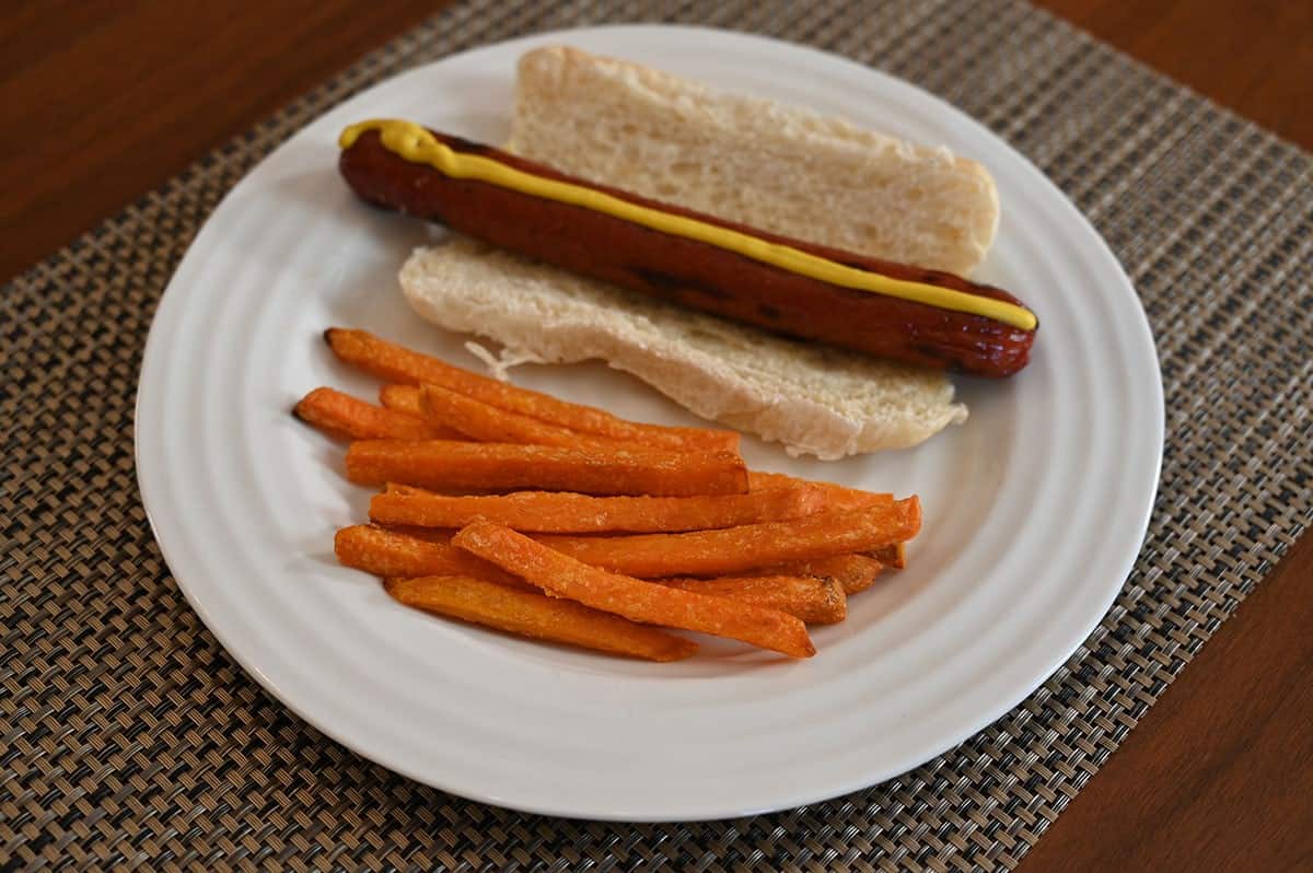 Image of cooked Costco Russet House Sweet Potato Fries on a white plate beside a prepared hot dog with mustard on it. 