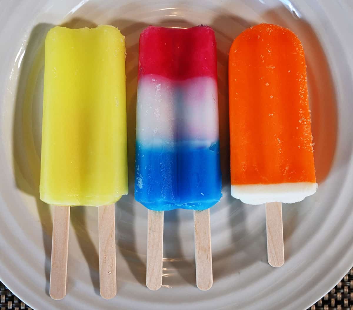 The three kinds of frozen bars on a white plate, banana, blue raspberry lemon cherry and orange creamsicle.