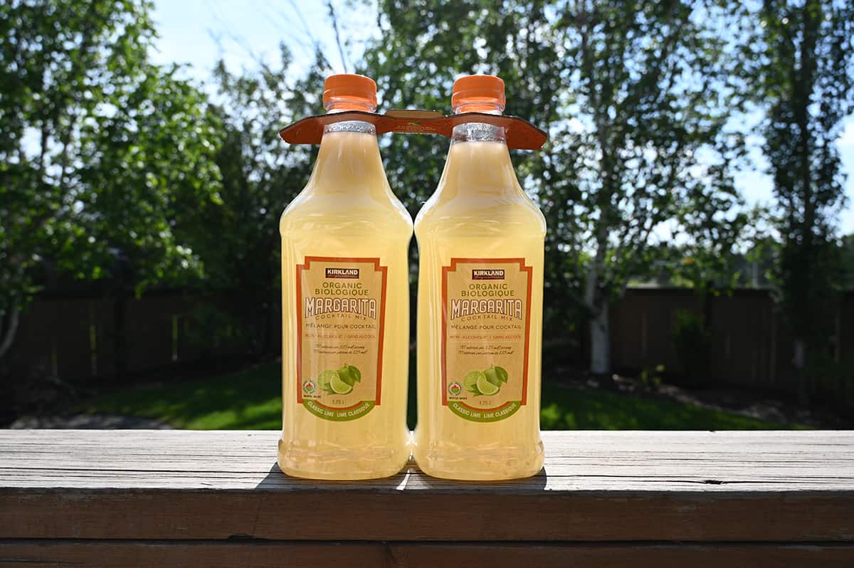 Image of the two-pack of organic margarita mix sitting on a deck rail with trees and grass in the background.