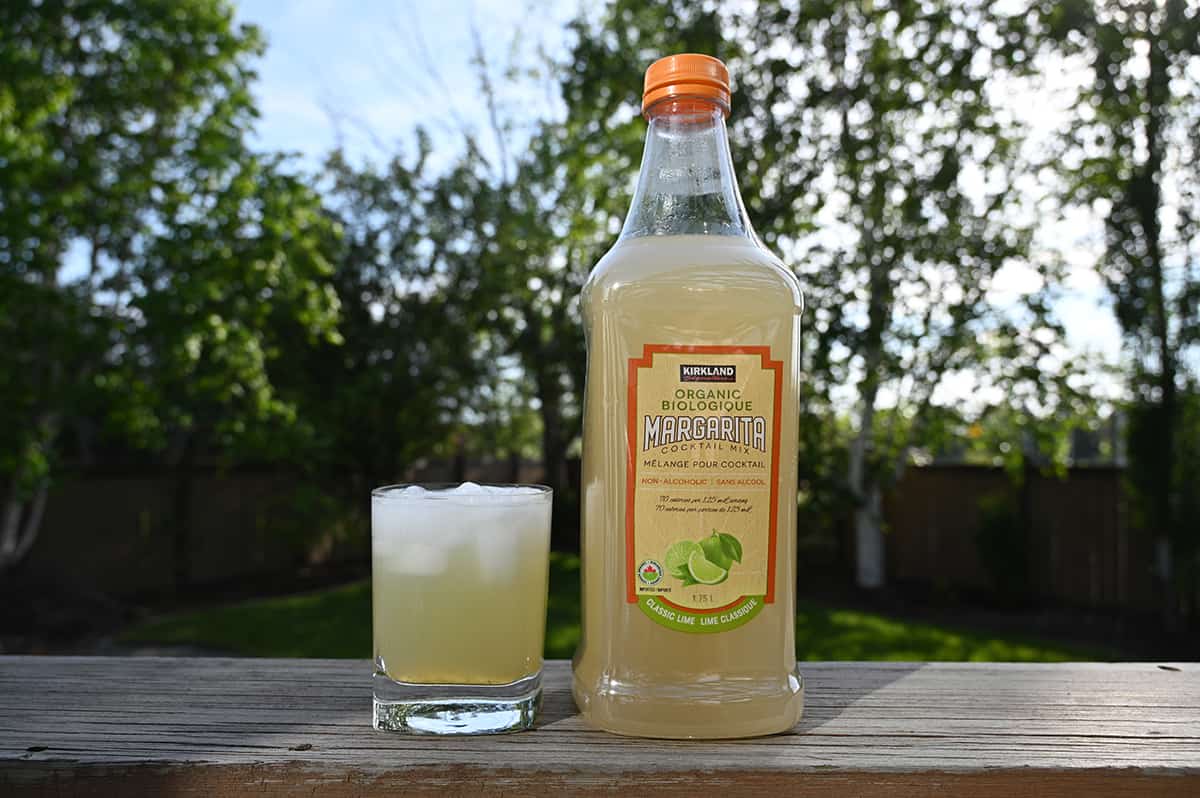 Image of the bottle of organic margarita mix beside a margarita cocktail in a clear glass with ice.