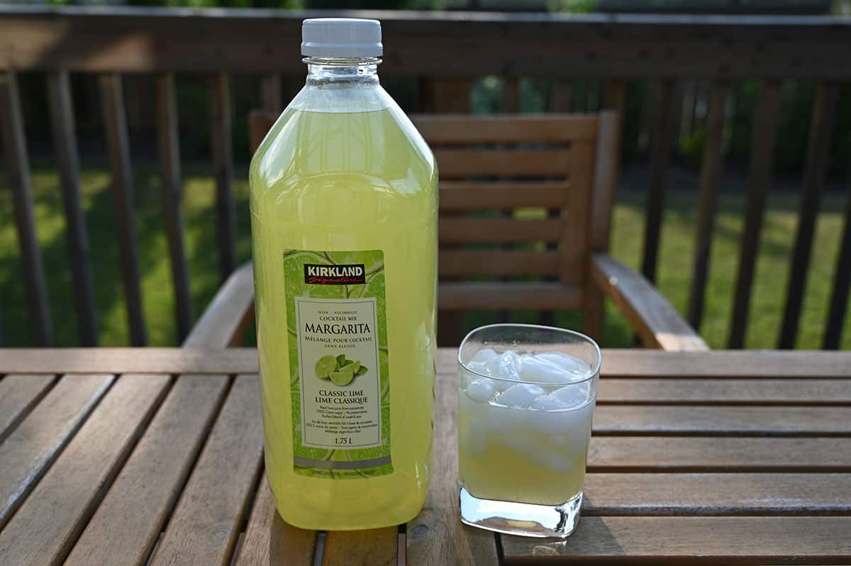 Bottle of the old margarita mix beside a glass of prepared margarita, on a table outside.