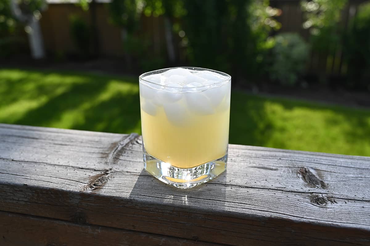 Image of a prepared margarita in a small glass with ice sitting on a wooden rail with grass and trees in the background.