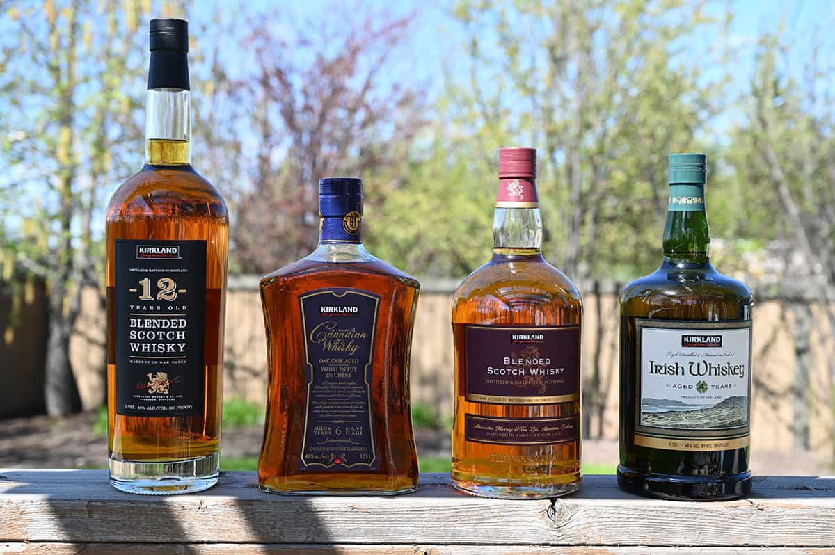 Image of four Kirkland Signature whiskeys from Costco lined up on a deck outside. From left to right, the bottles are the 12 Years Old Blended Scotch Whisky, Canadian Whisky, Blended Scotch Whisky and Irish Whiskey. 