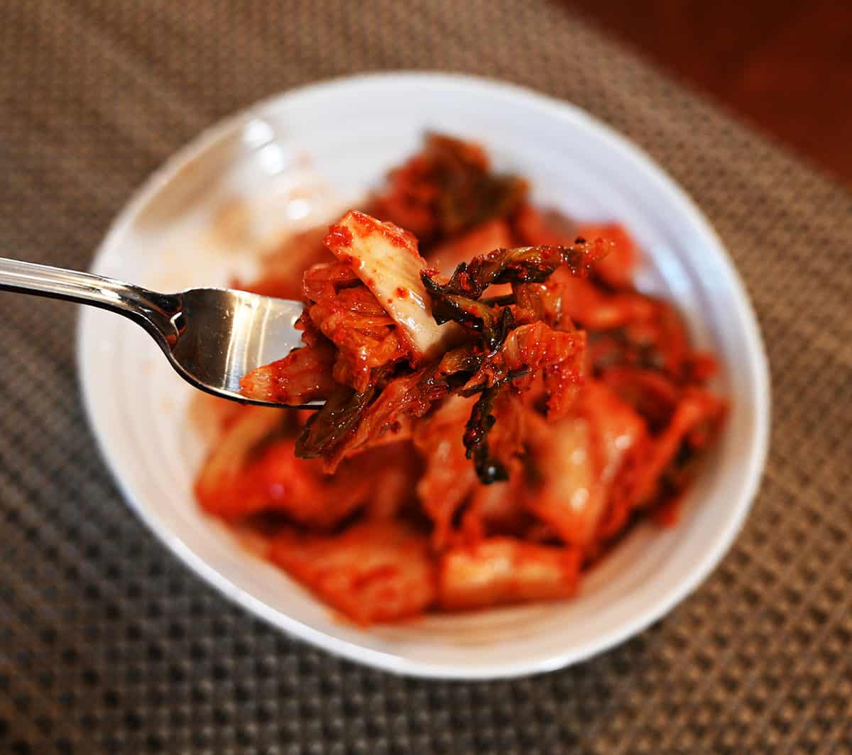 Closeup image of one forkful of kimchi with a white bowl full of kimchi in the background.