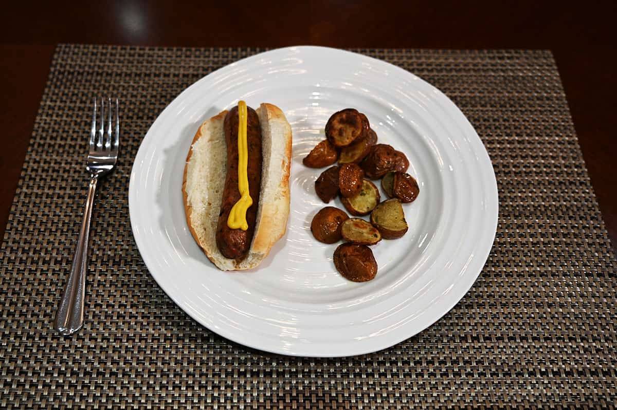 Image of a chicken sausage cooked and in a bun with mustard on it with a side of oven potatoes served on a white plate.