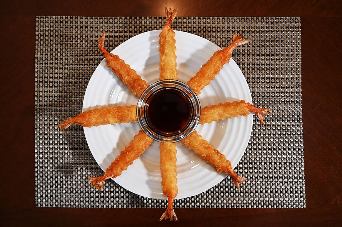 Top down image of a white plate with eight tempura shrimp on it and a bowl of dipping sauce in the middle.