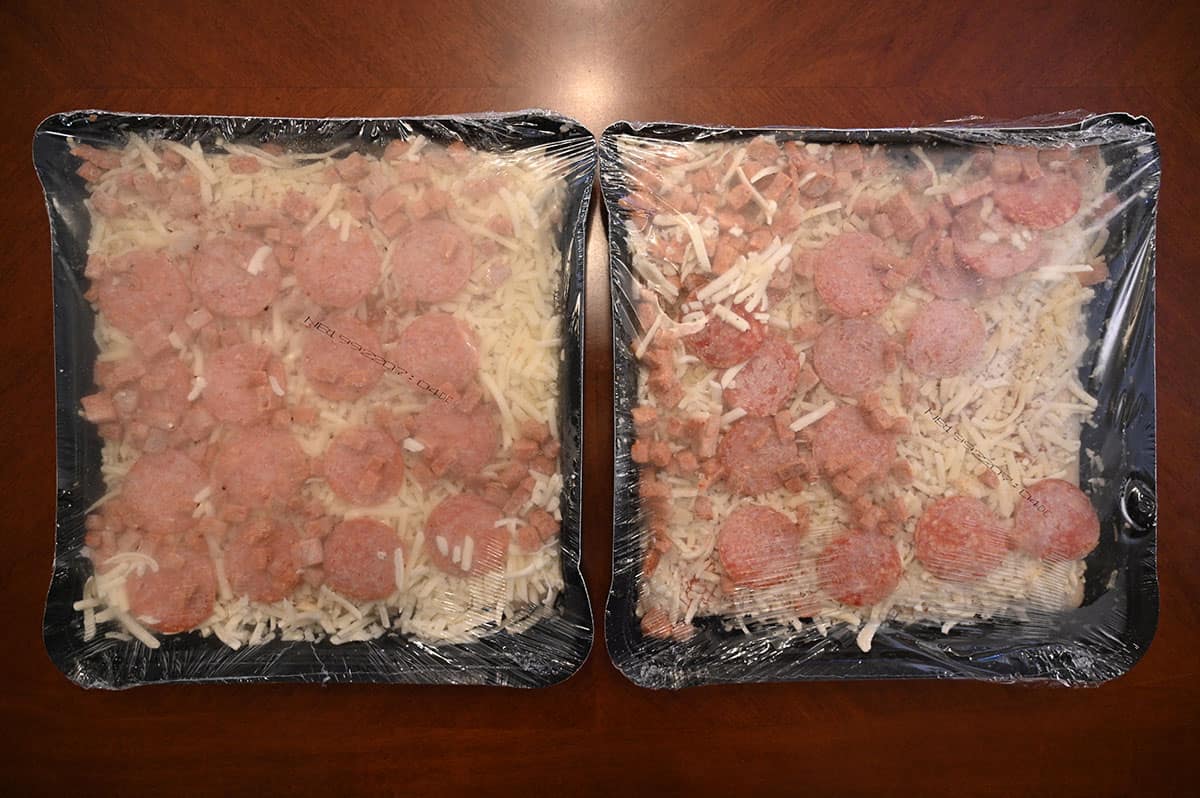 Image of the two frozen pizzas that come in the box with the plastic wrap still on, before baking.