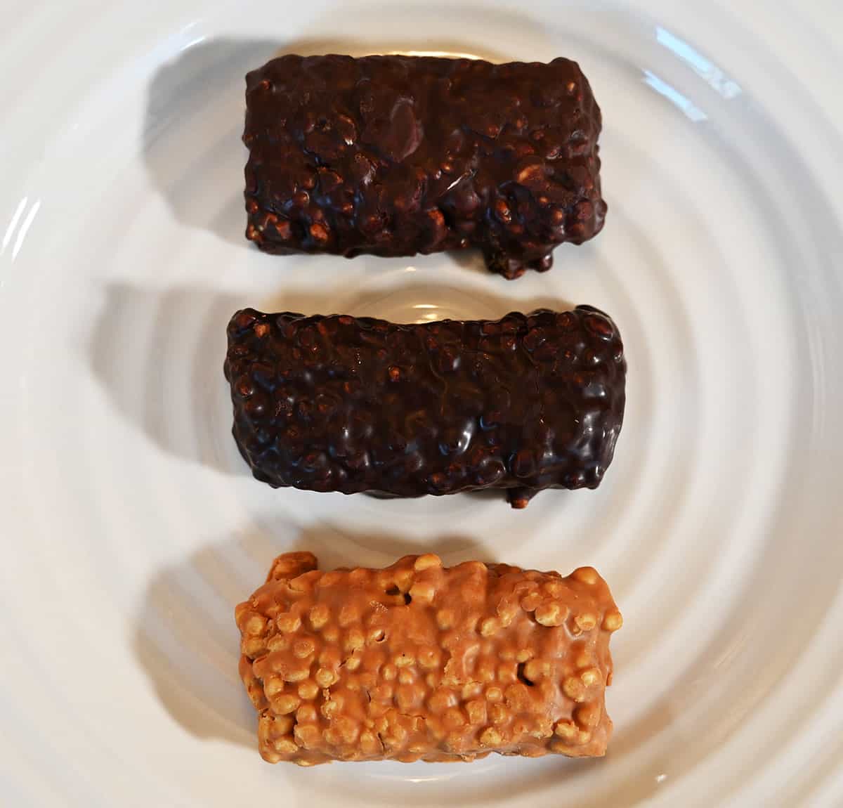 All three flavors of protein bars out of their packaging on a plate. Top to bottom, caramel almond, choco crisp and peanut butter.
