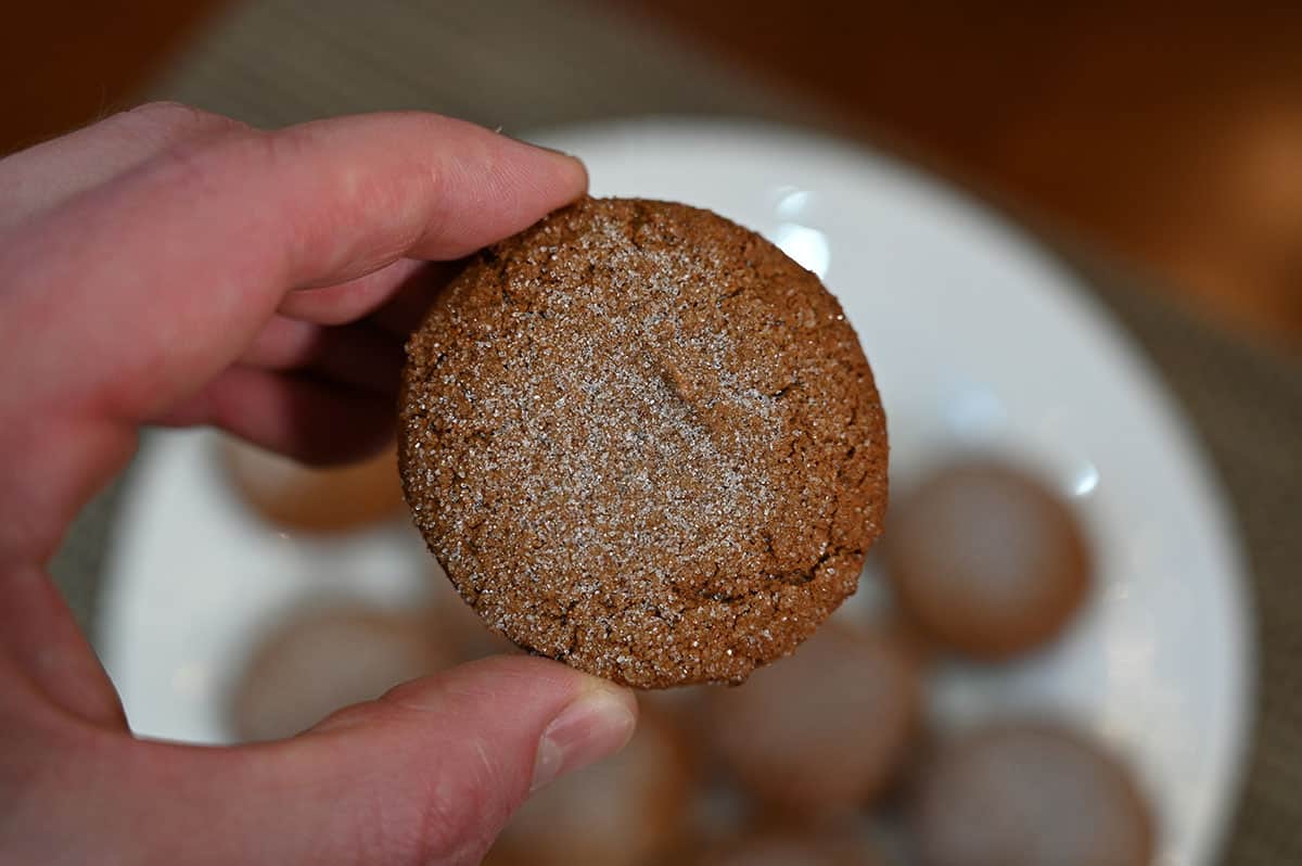 Closeup image of a hand holding one cookie so you can see the top of the cookie. Plate of cookies in the background of the image.