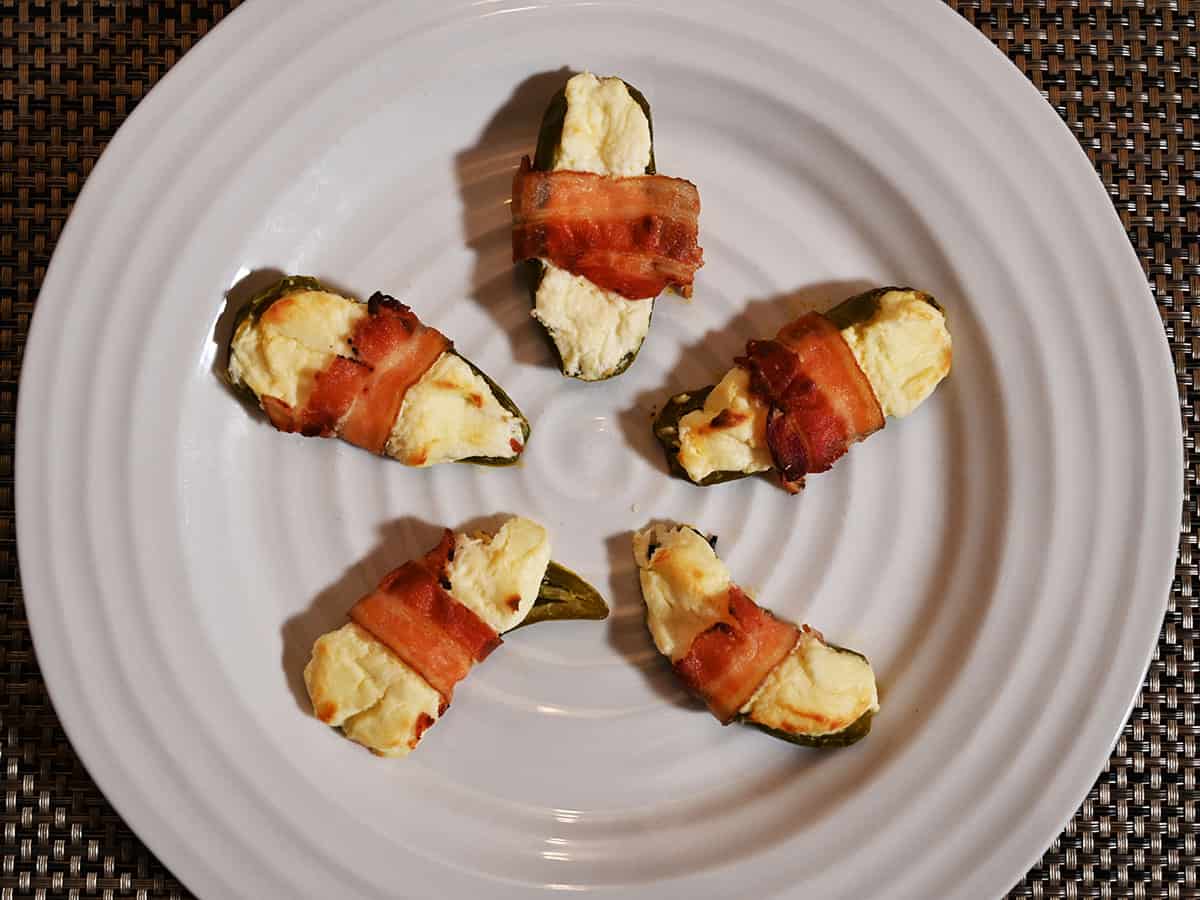 Image of five cooked bacon wrapped jalapenos on a white plate, served in a circle.