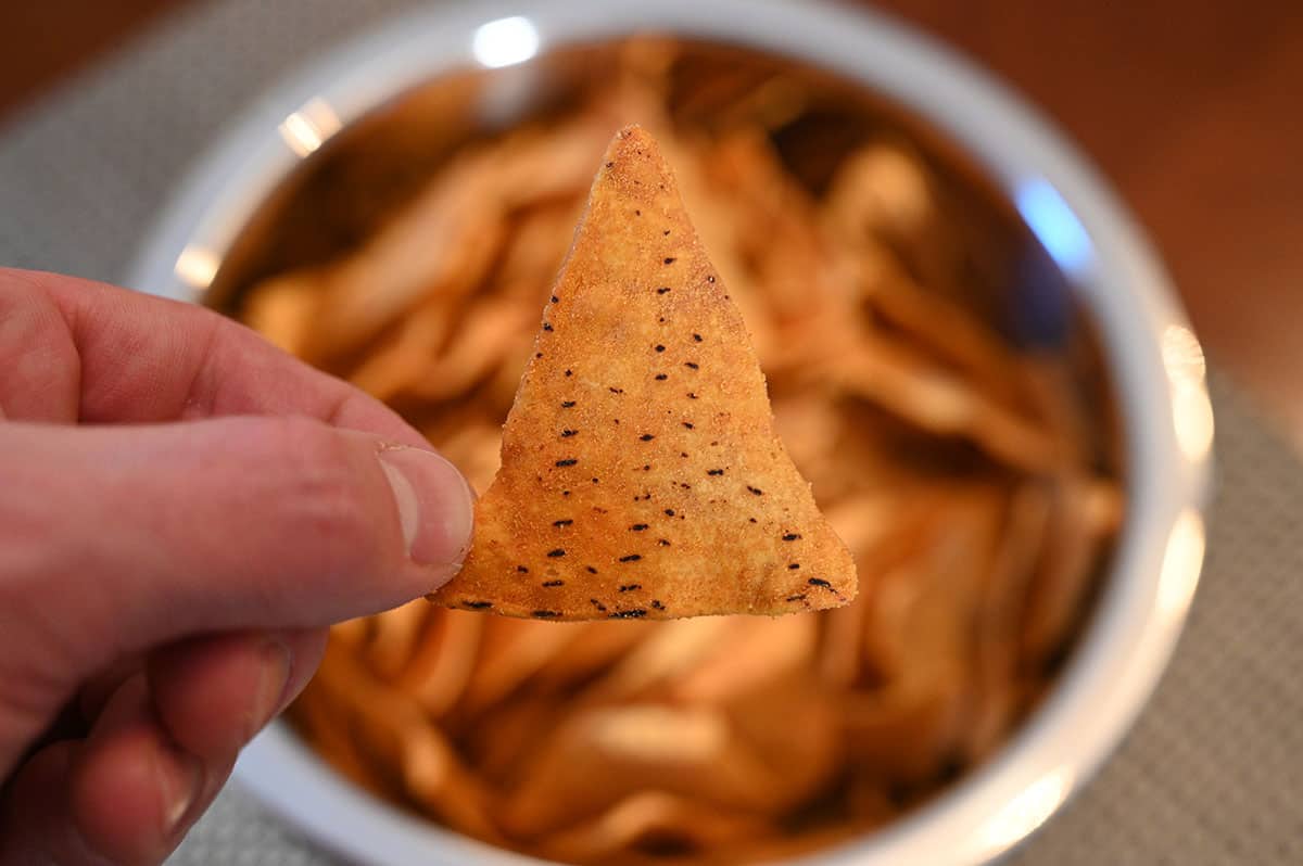 Image of a hand holding one nacho cheese chip with a bowl of chips in the background.