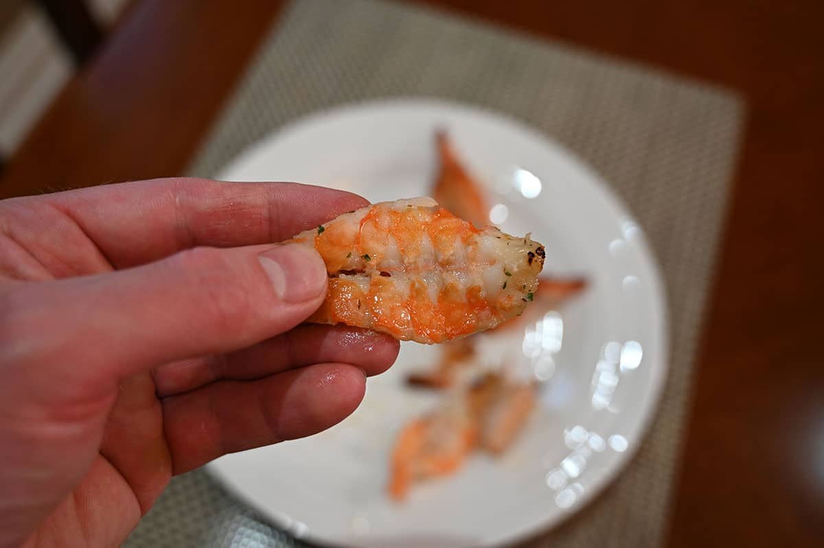 Closeup image of a hand holding one cooked butterfly shrimp with a plate of cooked shrimp in the background.