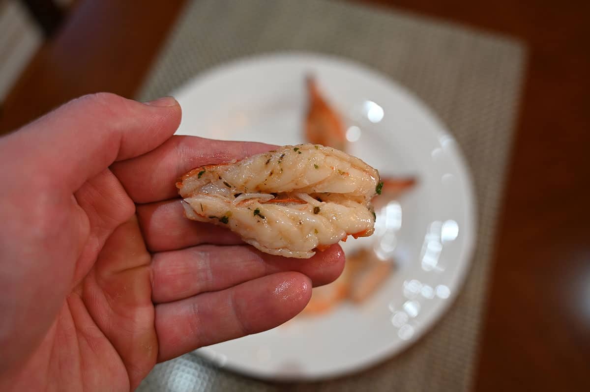 Closeup image of a hand holding one cooked butterfly shrimp without the shell on with a plate of cooked shrimp in the background.