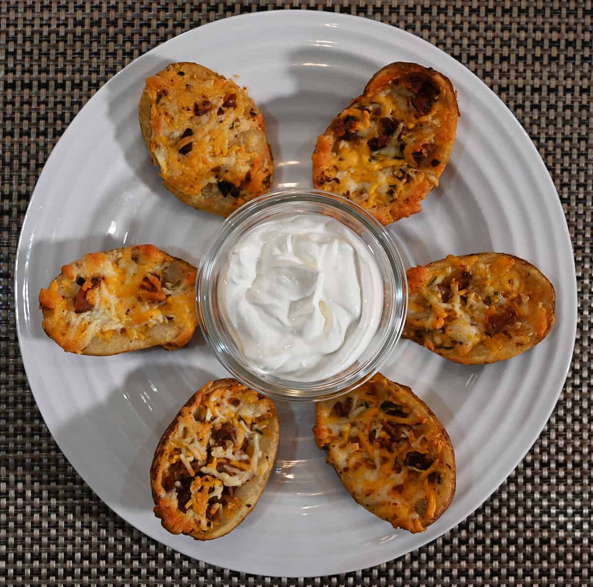 Top down image of six cooked potato skins on a white plate with a bowl of sour cream in the middle. 
