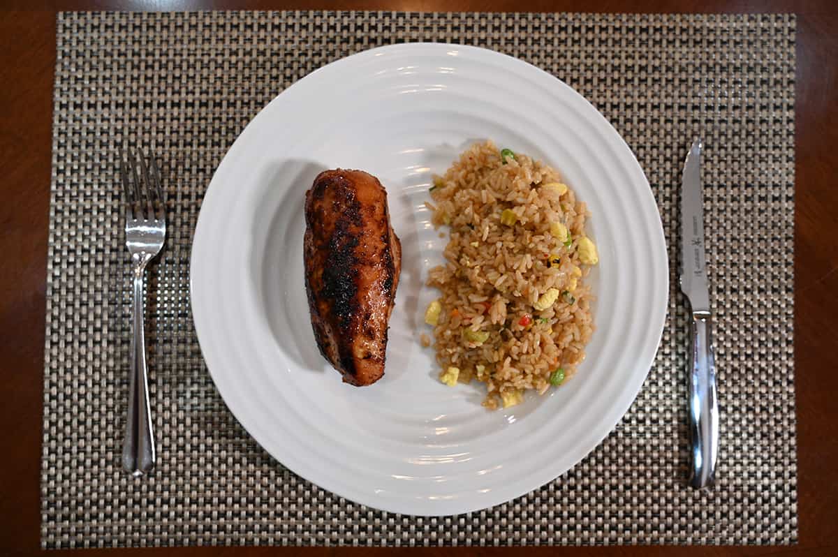 Top down image of a white plate with a fork and knife beside it, on the plate is vegetable fried rice and a chicken breast.