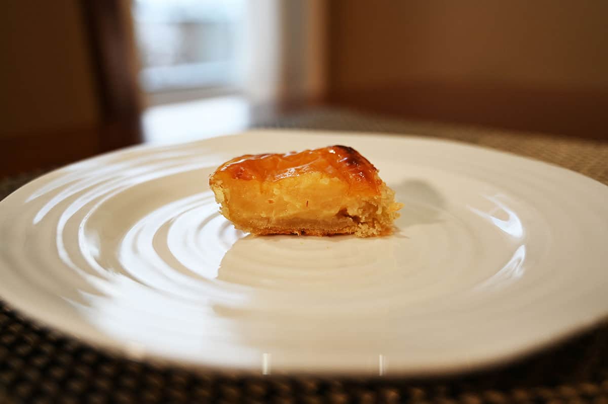 Side view image of a custard tart served on a white plate with a bite taken out of it so you can see the filling.