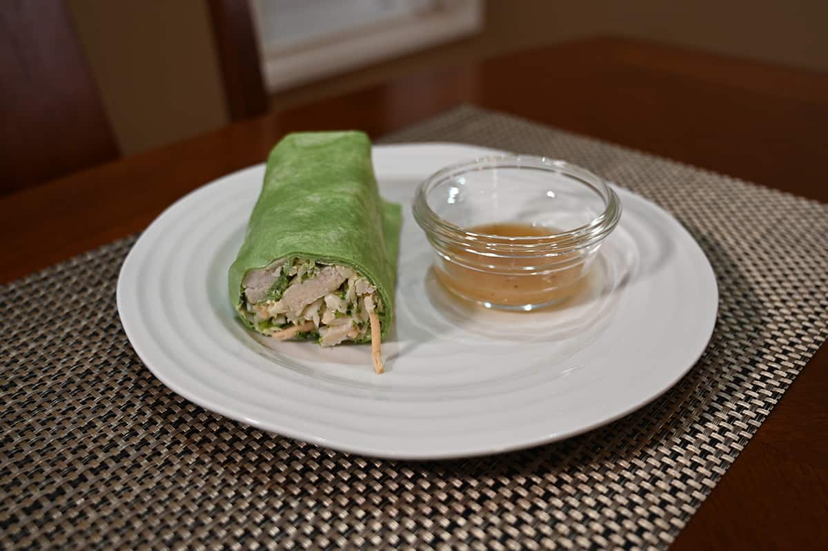 Side image of a wrap served on a white  plate with a bowl of sauce beside it.