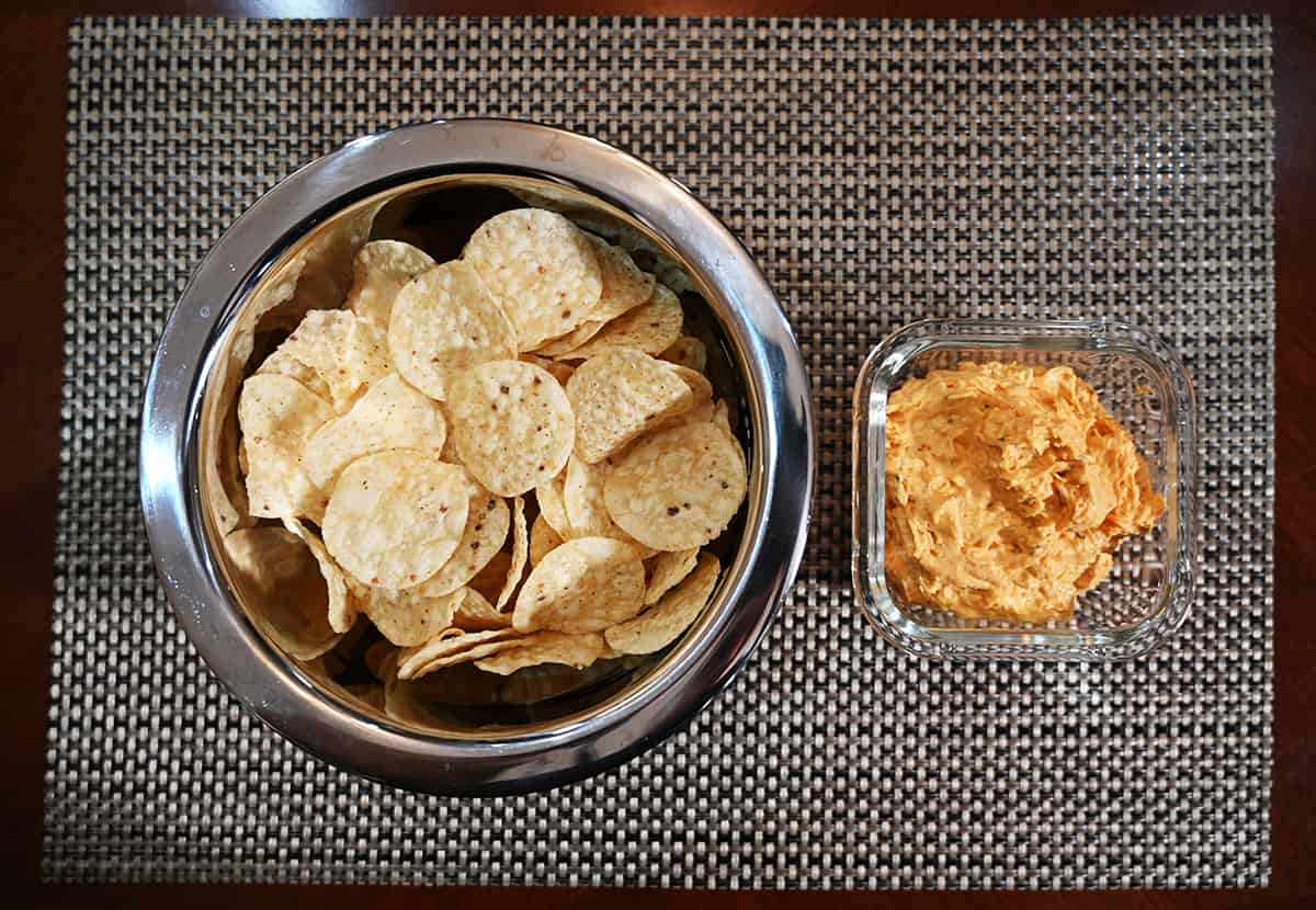 Top down image of the dip in a glass container beside a metal bowl with plain tortilla chips in it.