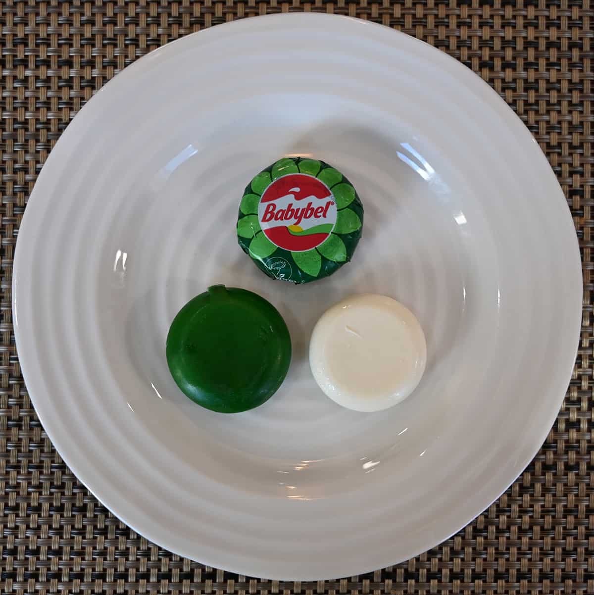 Image of three plant-based Babybel on a white plate, one unwrapped, one with the green wax seal still on and one in the wrapper.