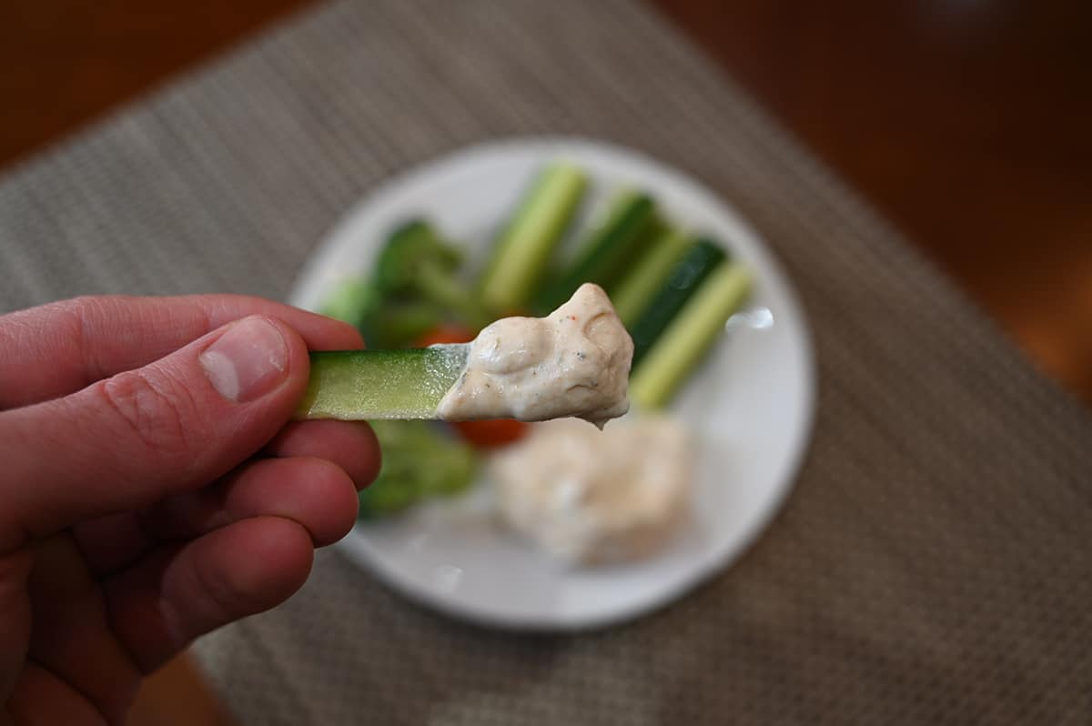 Closeup image of a hand holding a cucunber with dip on it in front of the camera with a plate of vegetables and dip in the background.