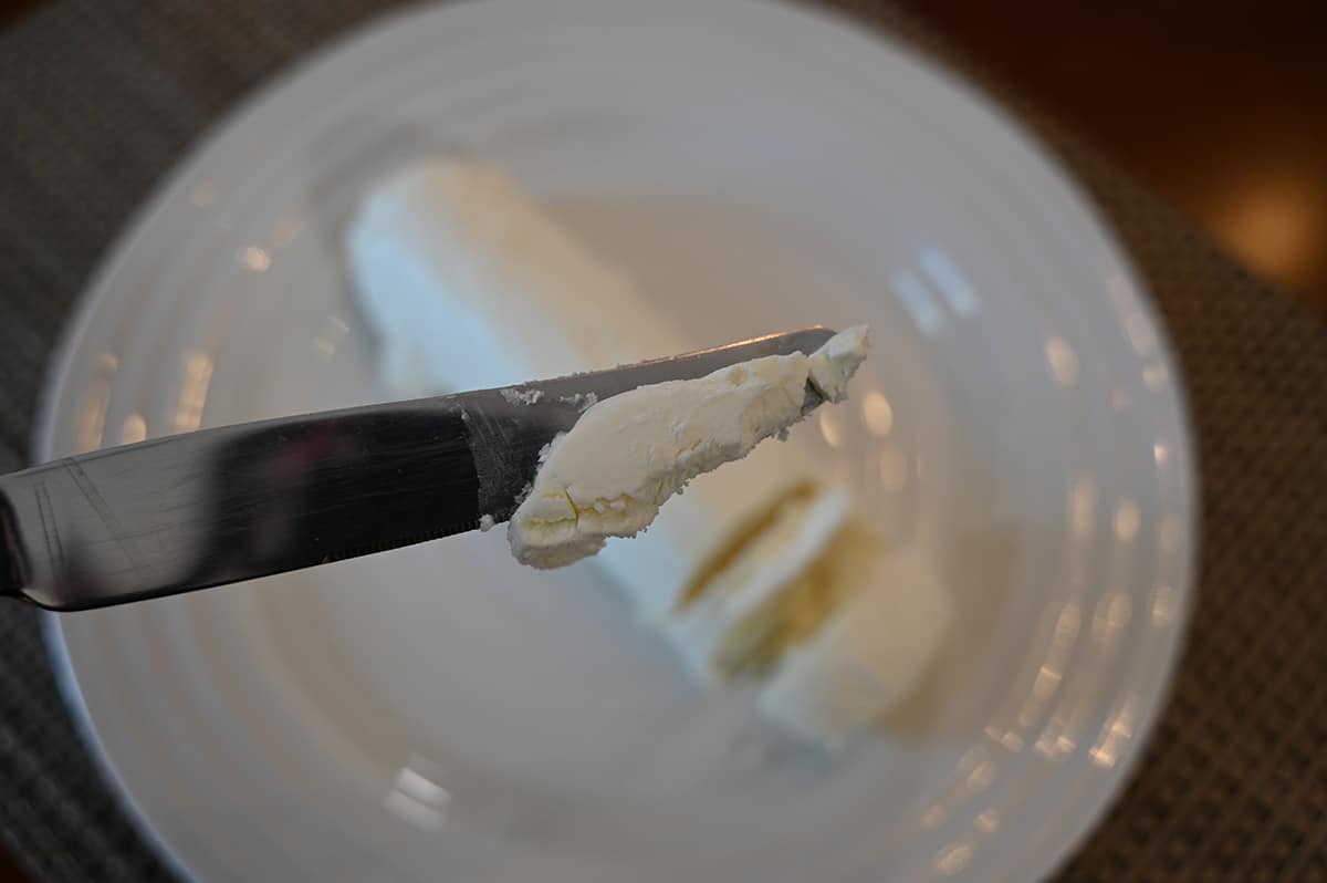 Closeup image of a knife with a spread of goat cheese on it with the plate of goat cheese in the background of the image.