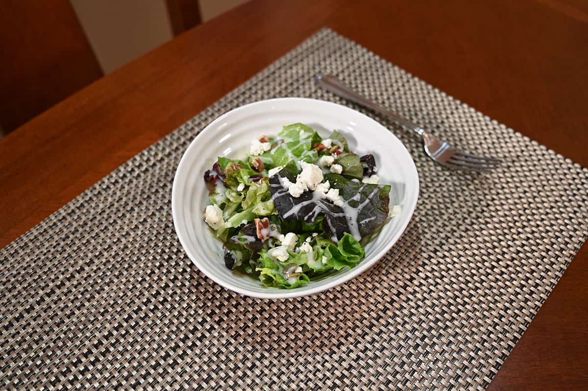 Side view image of a salad prepared with the goat cheese and a drizzle of dressing on top. Served in a white bowl with a fork beside the bowl.